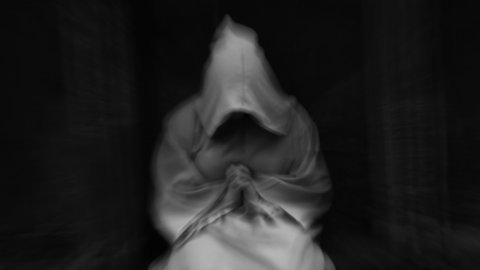 A blurry black and white photo of a hooded figure clasping its hands in a dark room.