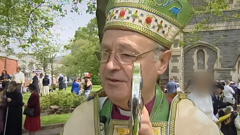 Former Bishop of Swansea and Brecon Anthony Pierce is a grey-haired man wearing thin-rimmed rectangular glasses. He is wearing a purple shirt with a dog collar and has green, white and gold robes.