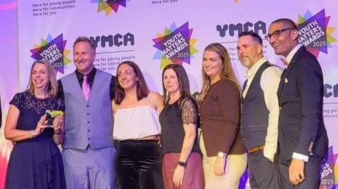 Seven people on a stage smiling at a camera off to the left of this image.