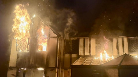 A large house that is on fire. Flames are soaring out of a window beneath a gable on the house, while flames are also shown spreading across both the lower and upper floors of the property. The yellow flames stand out against the blackness of night in the background.