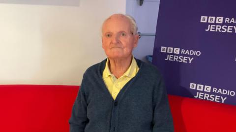An old man with grey hair and a bald spot on top, wearing a yellow shirt and blue cardigan.