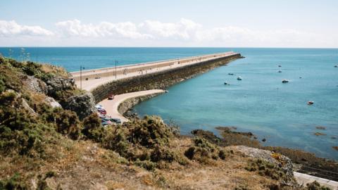 St Catherine's Breakwater stretches from the land in to the sea on a sunny day. There is coastal scrubland in the foreground and the bay is dotted with small motorboats. There are a few cars parked at the top of the slipway and people walking along the stone breakwater.