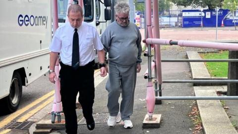 Anthonio Villafane is wearing a grey tracksuit with white trainers. He has grey/white curly hair and black glasses. He looks down as he is led by a Geoamey court officer who is wearing a white shirt, black trousers, black boots and a black tie. Anthony Villafane is handcuffed to the court officer by the wrist. 