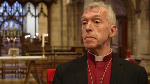 Andrew John in a red shirt and black blazer with a dog collar, looking at the camera in front of an altar. He has grey hair and brown eyes