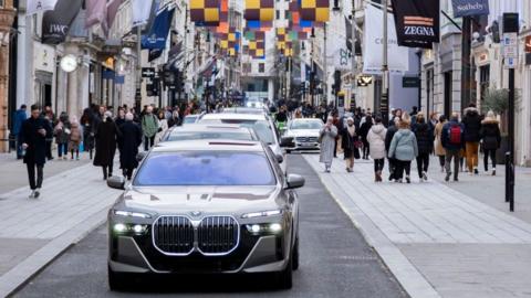 A luxury car drives along a shopping street in central London. There is a row of cars behind it. Pedestrians are walking on the pavement on both sides of the street.