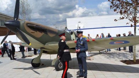 A historic military aircraft, painted in camouflage colors, stands outdoors on a paved area. Several people stand around the plane, including two individuals in formal military-style uniforms engaged in conversation near the wing.