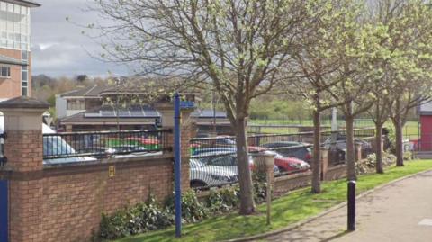 United Lodge can just be seen beyond the cricket club boundary wall which is edged with grass verge and trees. To to the left is the edge of the main cricket club building. A path runs to the front of the boundary wall.