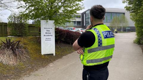 A police officer standing outside Birley Academy with his face turned towards the school building at the end of the drive.