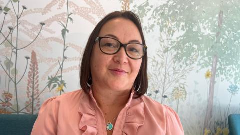 Tania Bartlett is sitting on a chair. She is wearing a pink blouse and has shoulder-length, brown hair. 