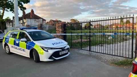 Police car outside gate leading to field - blue and white police tape is across the iron gate