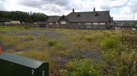 An overgrown desolate site filled with weeds in front of an old soot-stained school building.
