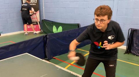Julie Norris is wearing glasses and has auburn hair.  She is looking intently at a green table tennis table as she hits a white ball. The ball is fuzzy as it is in motion.