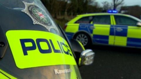 Close-up of a windscreen of a Police Service of Northern Ireland motorbike. The screen has the word police on it as well as the Police Service of Northern Ireland logo. In the background there is a police car. 