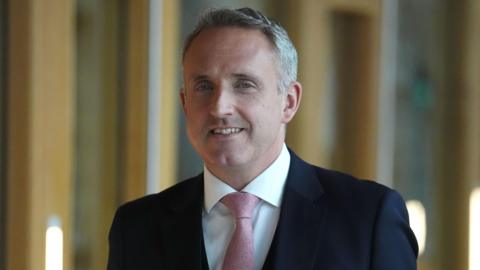 Alex Cole-Hamilton, wearing a navy suit and pink tie smiling as he walks in the Scottish Parliament.