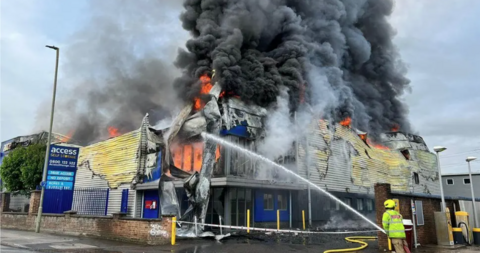 The former Access Self Storage facility in Byfleet, Surrey, while on fire in 2023. A firefighter can be seen fighting the flames with a big hose. Huge amounts of black smoke can be seen going up into the sky.