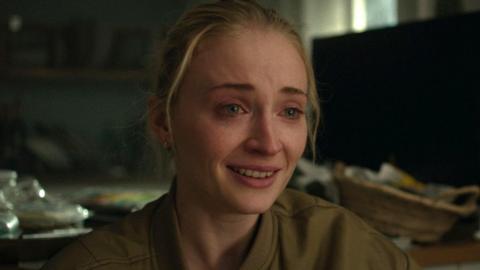 Picture of Sophie Turner looking upset, with tears in her eyes and her hair pulled back. She is wearing a khaki bomber jacket that is slightly unzipped. Messy blurred household items appear in the background.