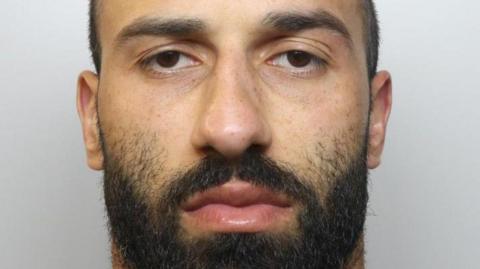 A mugshot of a young man with short hair and a dark beard. He is staring directly at the camera.
