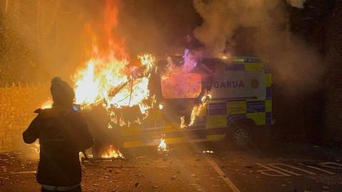 Police van on fire. Man stands in front.