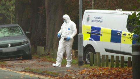 A forensic investigator wearing an all-white suit and mask is seen walking near a road with a police van in the background.