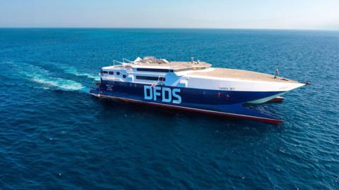 A blue and white catamaran ferry under sail on a clear day. It has DFDS printed in large white letters on the side and TARIFA JET in smaller blue lettering on the bow. 