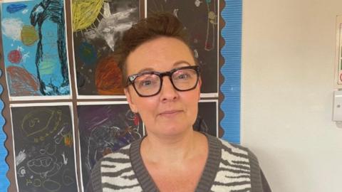 A woman with short brown hair, wearing black glasses, a grey and white jumper, stood in front of colourful pictures in a school