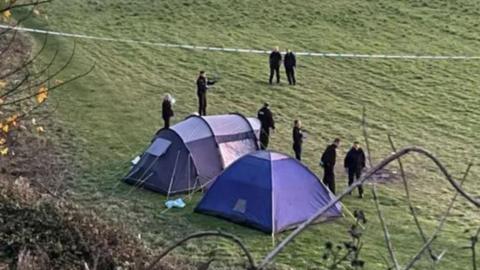 Aerial view of two blue tents in a field with a number of police officers standing around and blue police tape stretched across