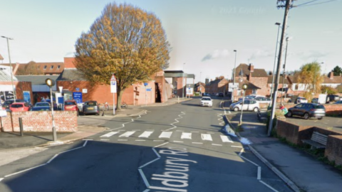 A pedestrian crossing on Oldbury Road in Tewkesbury. There are car parks on both sides of the road.