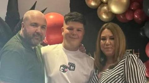 Dad Charlie and mum Victoria with their son in the middle wearing a white t-shirt. A balloon arch with gold and red balloons is visible behind them
