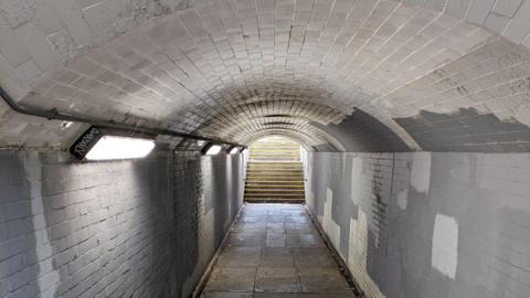 a dimly-lit, dirty-looking subway with steps leading out into the light outside. Graffiti is scrawled on the side one of three wall light strips. There are patches of white paint over the grey tiles on the walls. The ceiling is dirty and curved like in a tunnel.