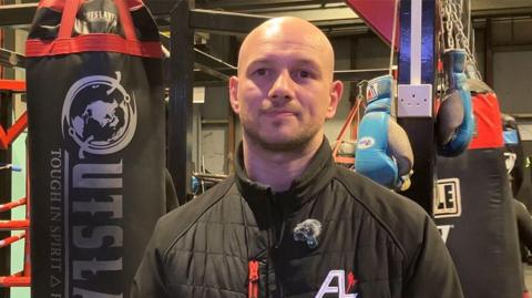 Head and shoulders of Adam Little standing in the gym with large black punching bags behind him. He is bald, with a very short brown beard and is wearing a black padded zip up jacket