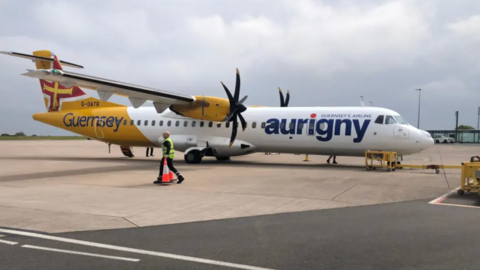 An Aurigny plane on the tarmac. It is white with a yellow back. It has the words Guernsey and Aurigny written on the side in blue writing.