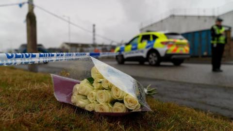 A bouquet of yellow roses placed on the ground with a police cordon in the background