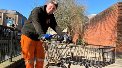 A man in a jumper and high-visibility trousers holding a trolley with gloved hands next to a river bank.