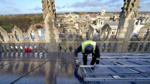 A photovoltaic engineer wearing a hard hat and yellow high-vis jacket at work on the installation of 438 new photovoltaic solar panels on the roof of the recently restored Chapel at King's College Cambridge. The panels will meet 100% of the energy needs of the Chapel and will reduce the College's carbon emissions by more than 27 tonnes each year.