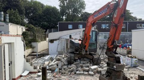 A small digger is parked on a large pile of pieces of concrete with its bucket resting on the ground. A building to the left has been partly dismantled. Behind the digger is a two-storey building.