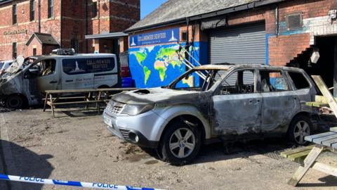 The remains of a Collingwood Seafood-branded van stands outside the shop following a fire. A similarly destroyed car has been abandoned outside the shop. with its rear facing the building. The wall behind it has been knocked down. The building is a single-storey brick construction with a large, central metal garage-style shutter and a pitched, tiled roof. There is a mural to the left of the shutter depicting a map of the world, below a blue sign showing the company name. A line of blue and white police tape is in the foreground.