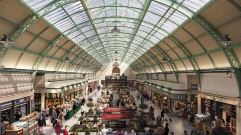 An elevated view of the Grainger Market, looking down at an arcade featuring a range of stalls selling plant, fruit and veg, as well as cafes and bakeries. Above is the barrelled glass roof.