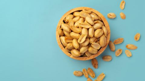 Bowl of peanuts on a duck-egg blue background, there are multiple peanuts outside of the bowl, suggesting they have been spilled