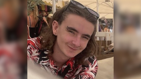 Jamie Collins wearing a black, red and white patterned shirt and sunglasses on his head. He has shoulder length brown hair and piercings in his ears, eyebrow and nose. He appears to be sitting underneath a white marquee on a sunny day.