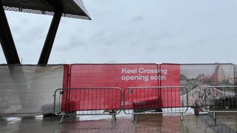 A red and white banner reading Keel Crossing opening soon covers a fencing in front the footbridge.