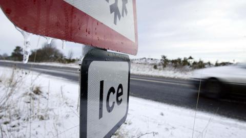 A stock image of a road sign warning of ice in the foreground, with a speeding car on a snowy road behind it.