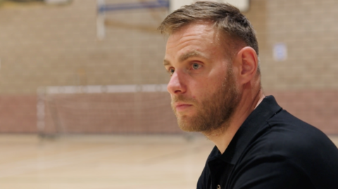 A head and shoulders profile view of a man with short straight brown hair and a beard, wearing a black top, inside a leisure centre, with brick walls and goal mouth in the background.