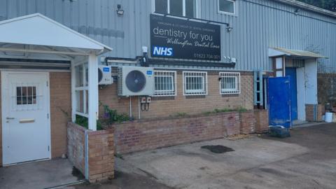 The entrance to Wellington Dental Care on the Lowmoor Industrial Estate in Wellington. The building is made of bricks and corrugated iron. The lower windows are small and have white iron bars across them and the building has two entrances, one with a white door and another with a blue door.