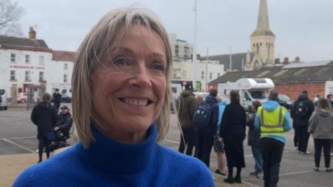 Jill Douglas wearing a blue turtleneck jumper. She has short blonde hair and is smiling. There is a group of people behind her standing in a car park.