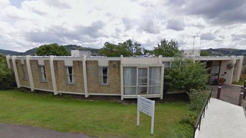 Wyatt House in Stroud. It is a curved, single-storey, brick building, with large white windows at the front. There is a lawn in front of it.