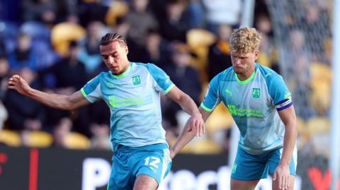 A photo of Cameron McGeehan and Nesta Guinness-Walker playing football in their light blue and fluorescent green Northampton Town third kit in March