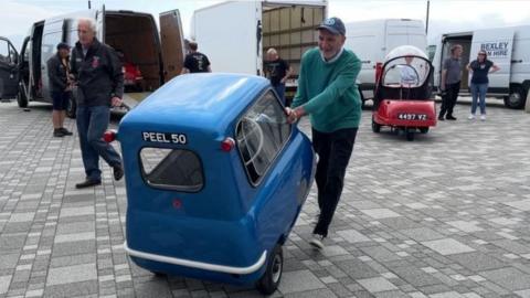 A man in a green jumper wheels a blue P50 through a car park.