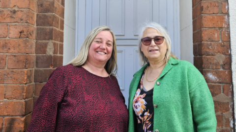 A woman with blonde hair and a dark red top beside a woman with grey hair, sunglasses and a green top in front of a white house door