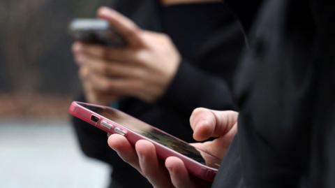 A person holds a pink mobile phone in their hand. In the background there is a second person also holding a mobile phone. 
