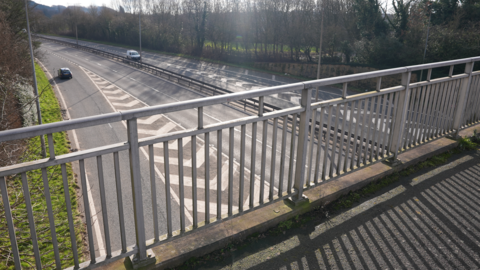 The bridge has a metal railing fence either side of it as it spans the A442. The carriageways can be seen below with a vehicles on each side.